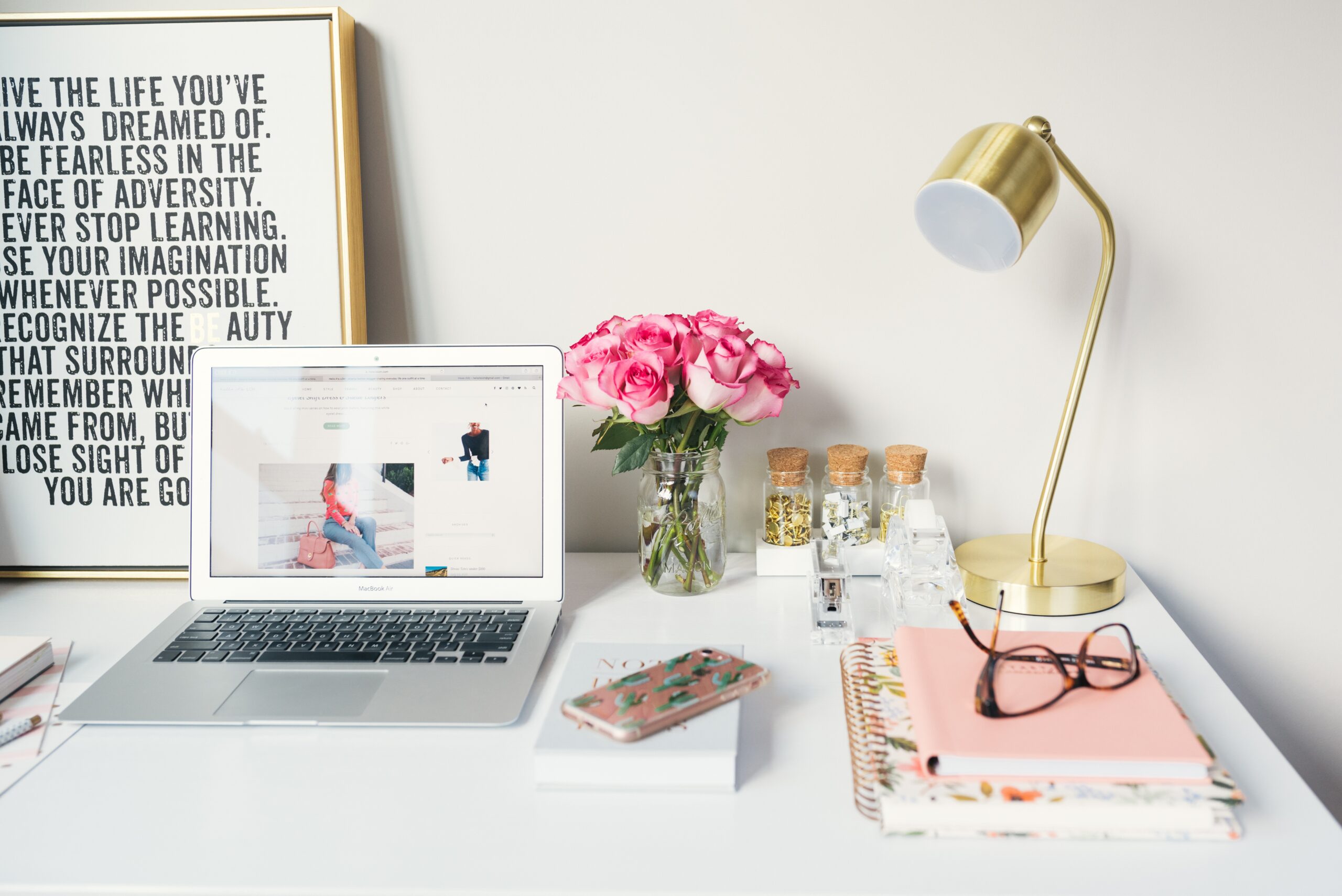 A copywriter's desk covered in delicate details like a brass desk lamp, open laptop, pink flowers and notebooks, and a motivational text poster in the background.