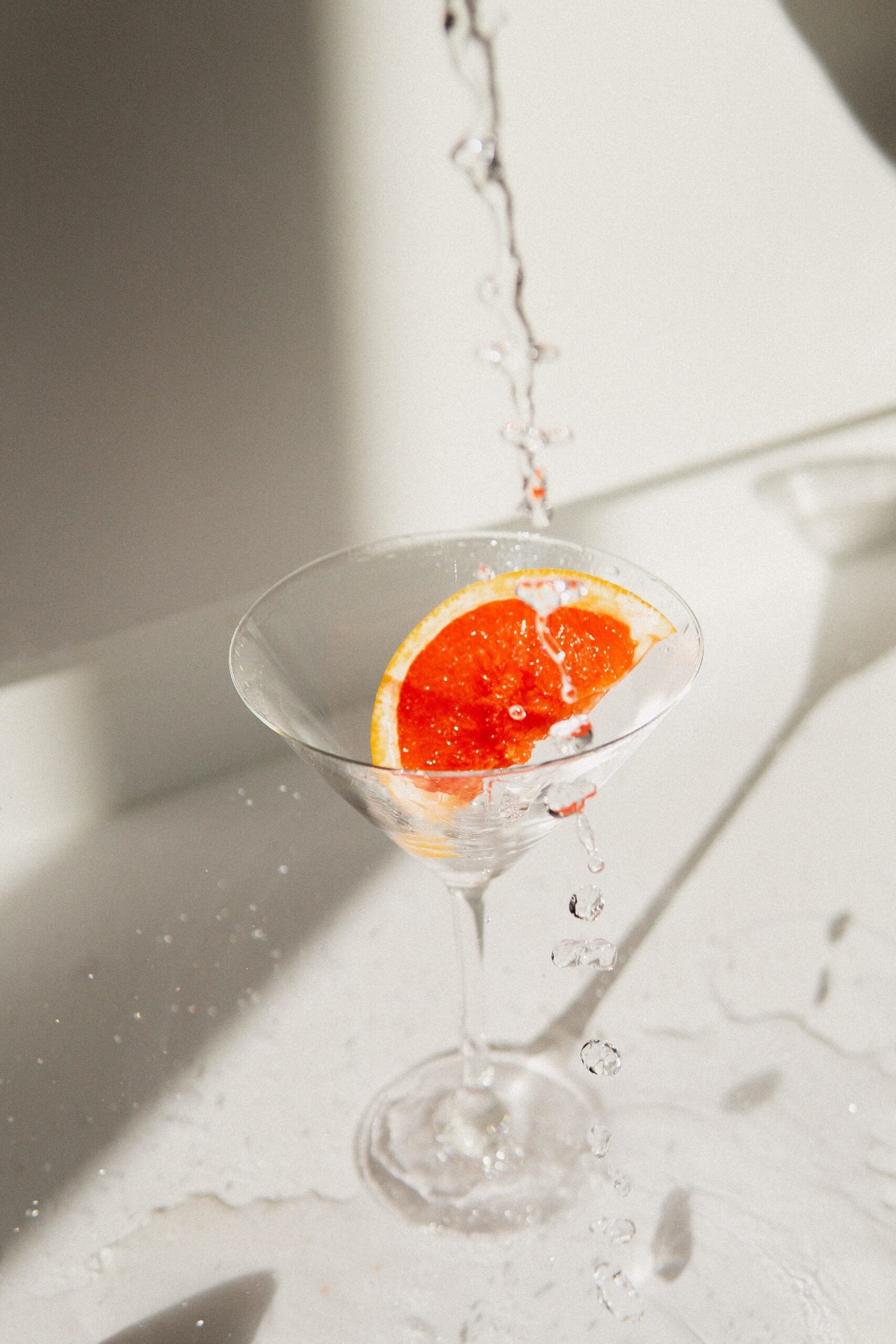 Liquid pouring into a martini glass with a slice of grapefruit in it and cascading over and out of the glass, splashing on the counter in a way that would grab your attention.
